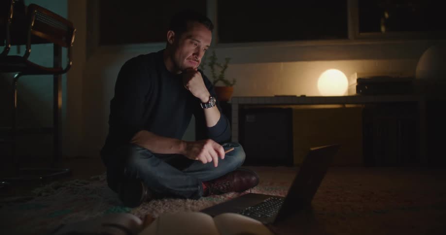 A man plays with a pencil while thinking then types on a laptop while sitting cross legged on the floor