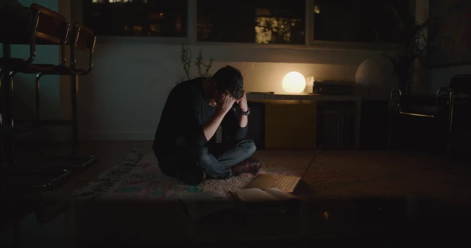 Tired man with sweatshirt and jeans sits on floor closing laptop in dark room