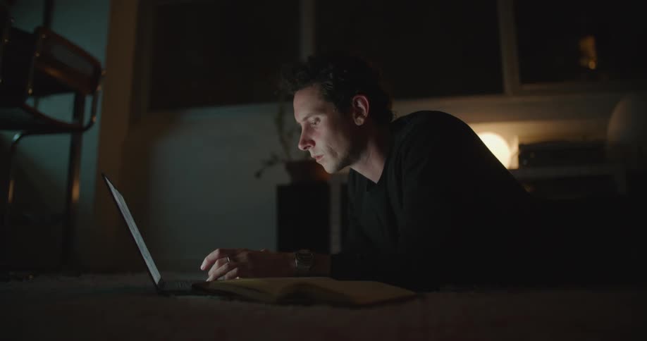 A man is lying on the floor of a dimly lit room and working on a laptop computer as he looks at a book