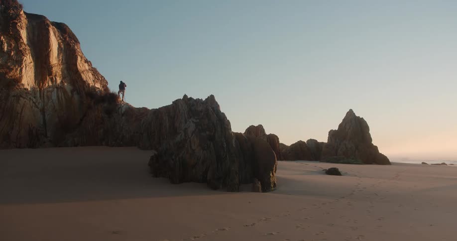 A man is standing on a rock formation at a beach as sunlight shines from a clear blue sky
