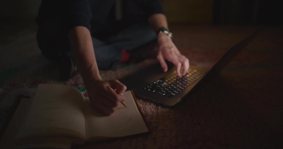 A man takes notes in a notebook while working on his laptop at home