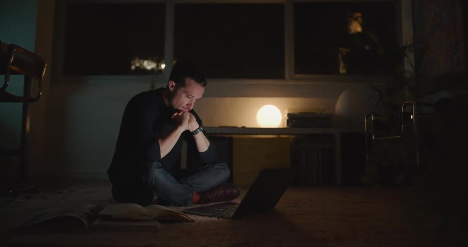 Tired and bored man uses laptop sitting on floor next to dining table