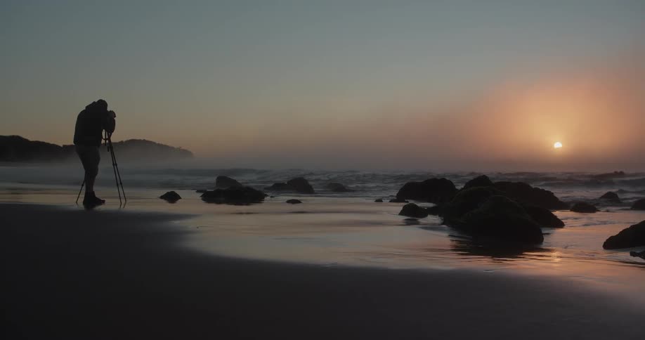 A man is standing on the beach with a camera on a tripod as waves roll into shore and the sun sets in a hazy sky in the distance