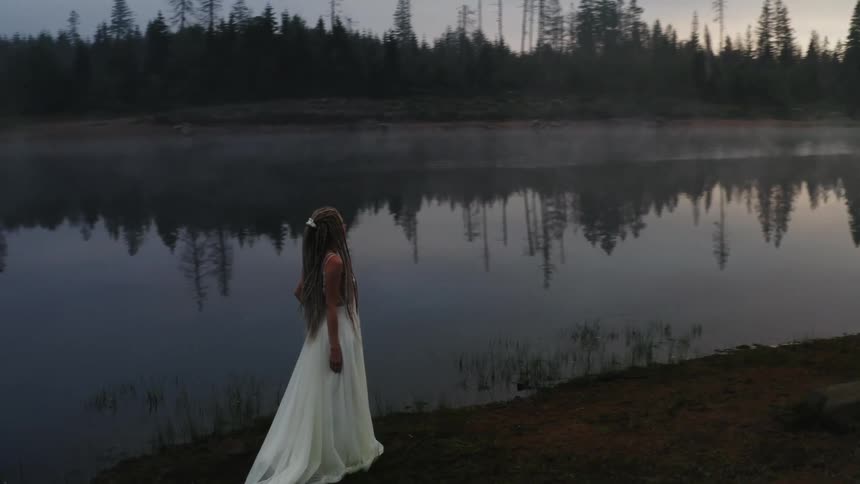 A woman in a long white dress stands next to a very still lake in the forest