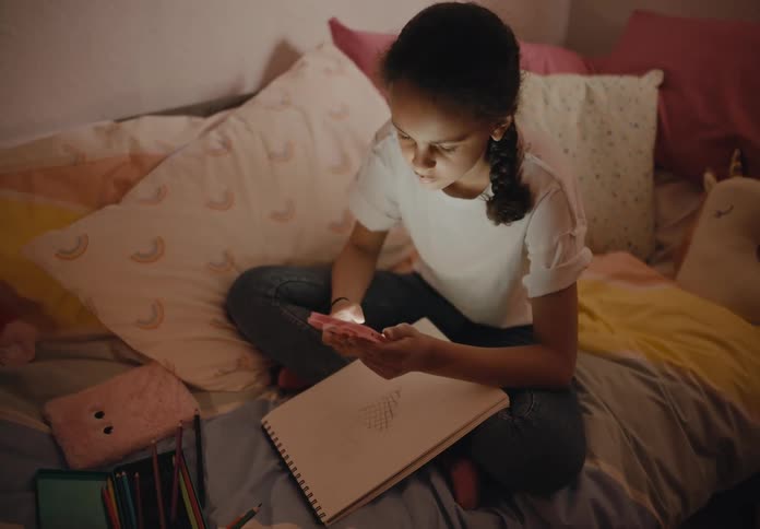 A girl is sitting on her bed doing homework and she stops to play with her phone