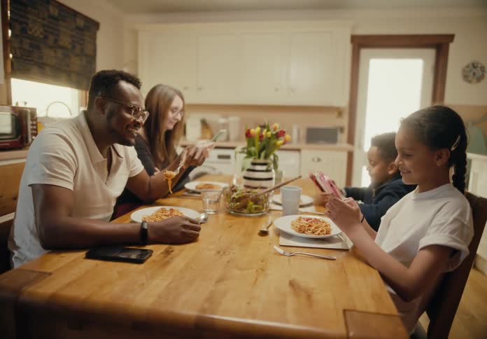 A family is sitting at a table in the kitchen eating dinner and looking at their phones