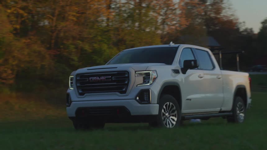 A person drives a white pickup on the grass field near a farmhouse