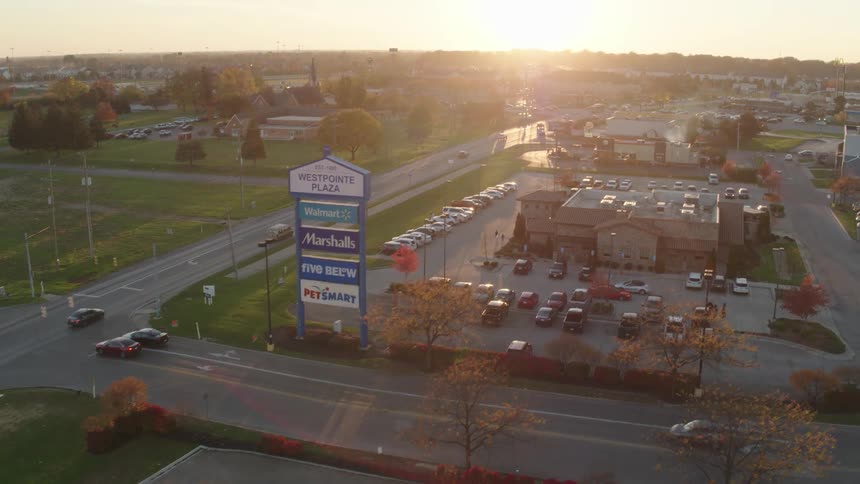 Traffic moves on a street past a shopping plaza and cars fill the parking lot as the sun shines brightly on the horizon