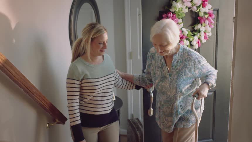 Young woman helps an elderly lady with a cane climb a staircase at home