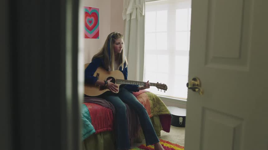 A teenage girl is sitting on her bed playing an acoustic guitar