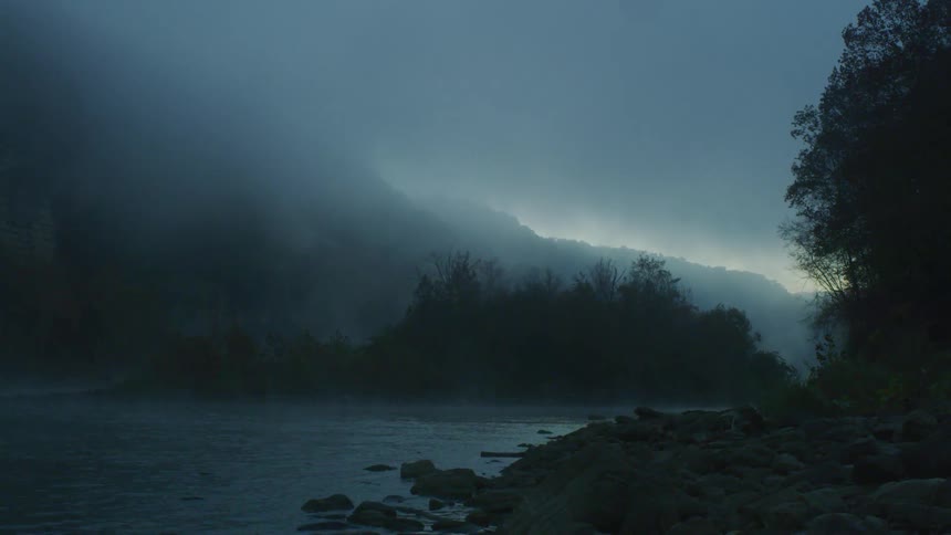 Gray clouds and fog over a river and hill as raindrops hit the water surface