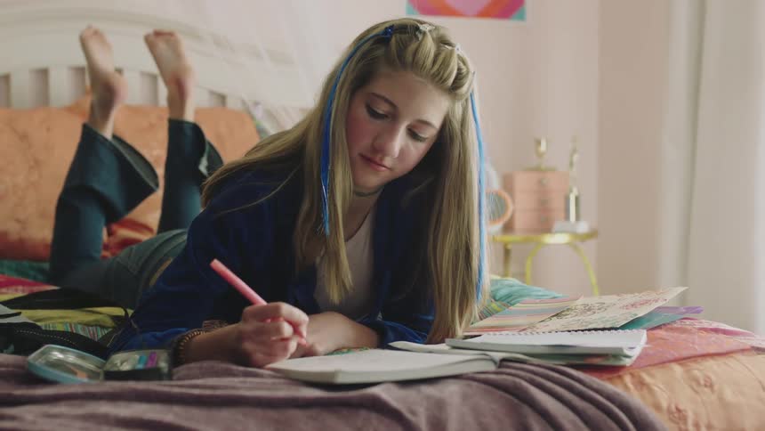 A teenage girl is lying on her bed and writing in a notebook with a pink marker