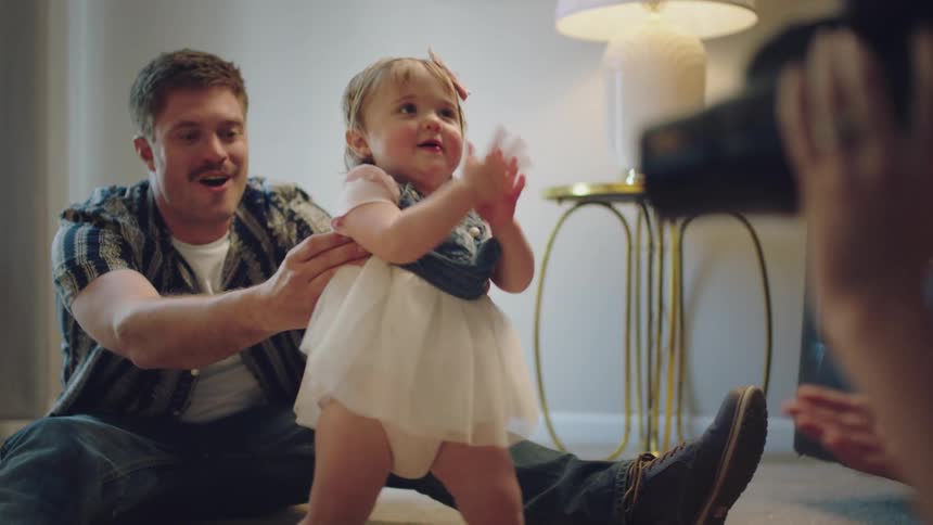 Dad holds onto his little girl as she takes her first steps toward mom who is filming