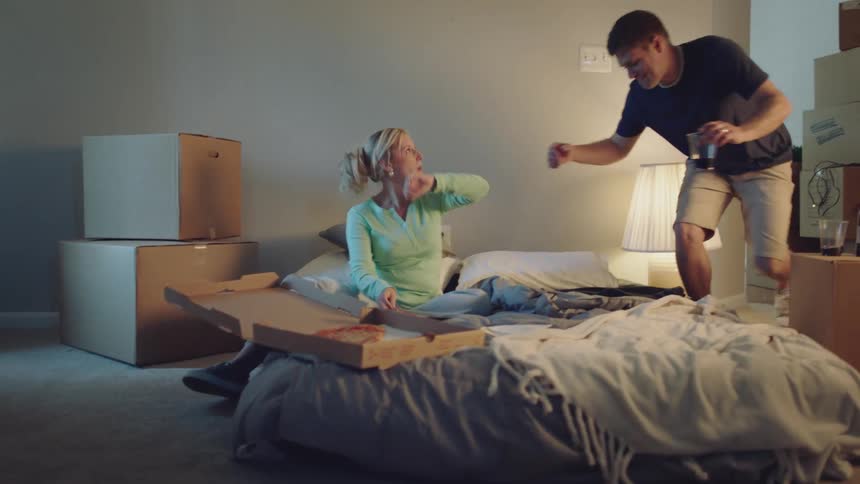 A man and woman are sitting on a mattress on the floor eating pizza and drinking wine having a good time