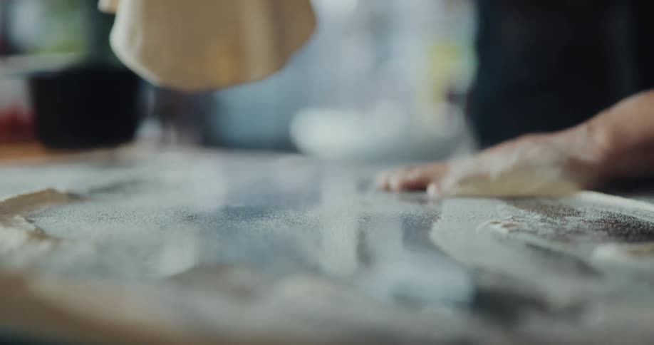A cook is covering flat round dough in flour on a table and they pick up the dough and push away the flour