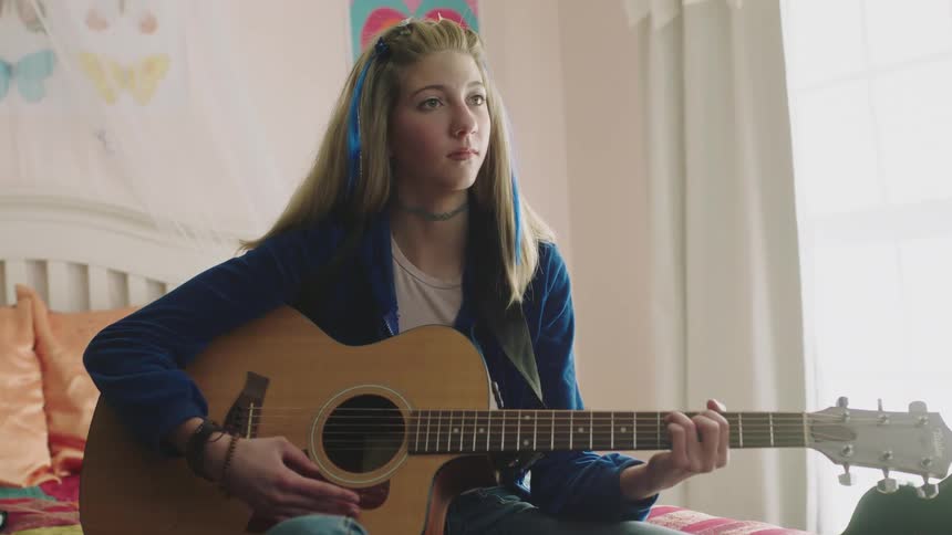 A teen girl sitting on her bed playing the guitar pausing as she thinks