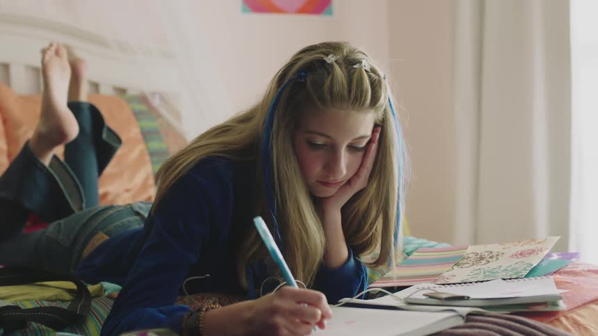 Teenager lies on her stomach on the bed listening to music and writing in a notebook