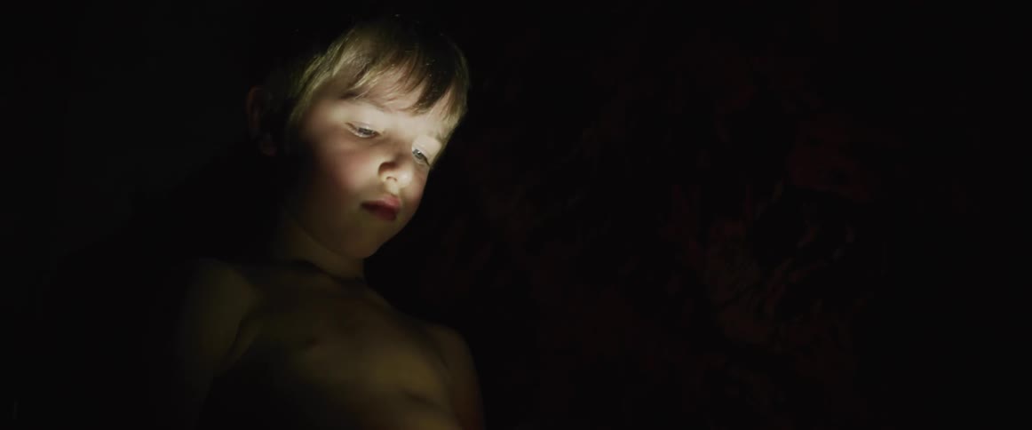 A little boy is standing in the darkness as the light from a flashlight shines on his face