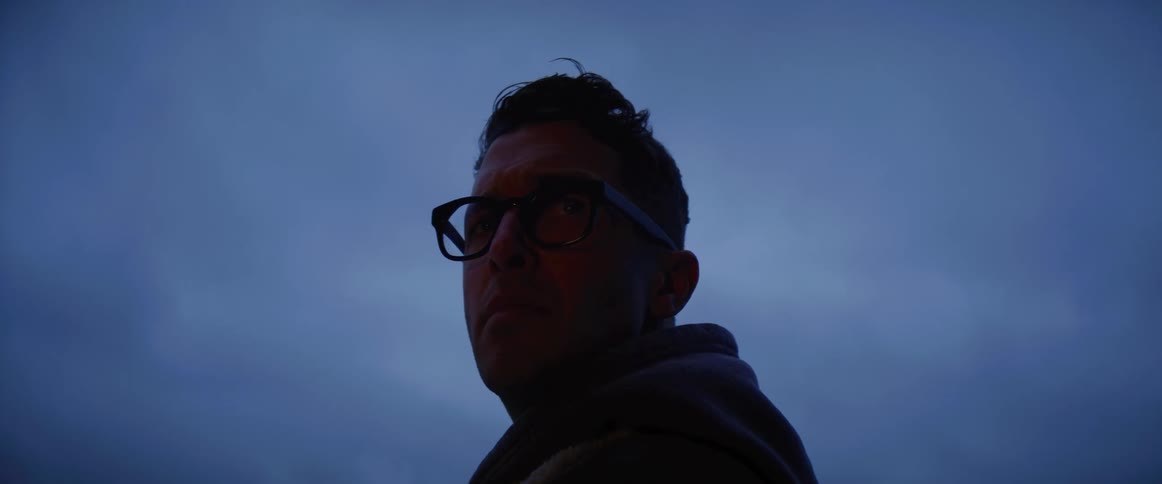 A man wearing glasses and a coat is standing outside and as he turns around and looks ahead a shadow moves over him