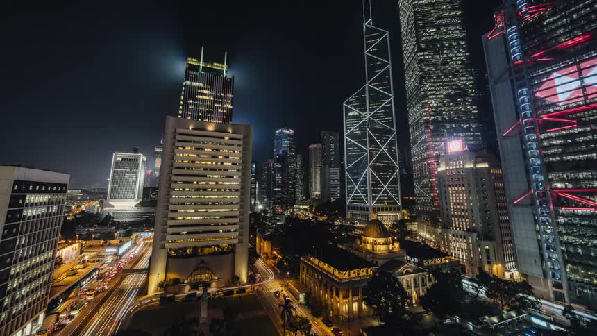 Traffic moves around a large city and the lights on tall buildings and skyscrapers glow as day turns to night