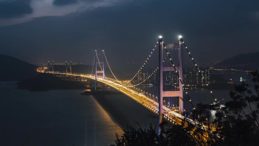 Water vessels cross under busy suspension bridge near city downtown as night falls