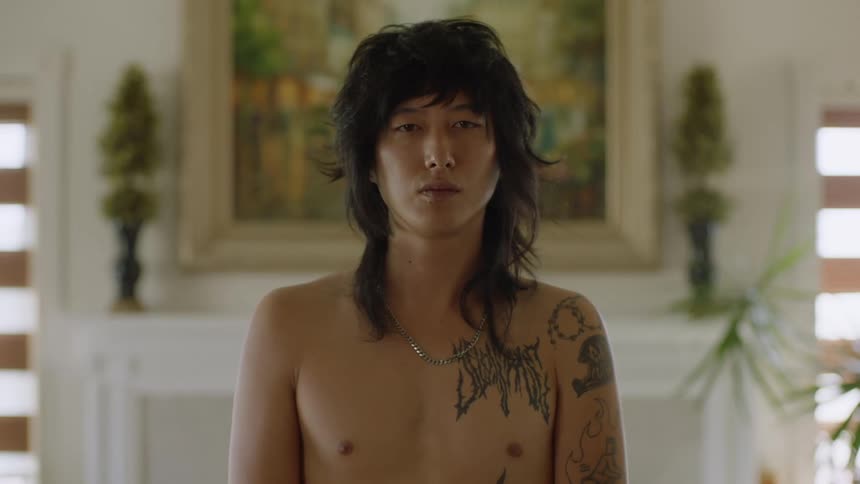 A tattooed man poses bare chested in front of a painting