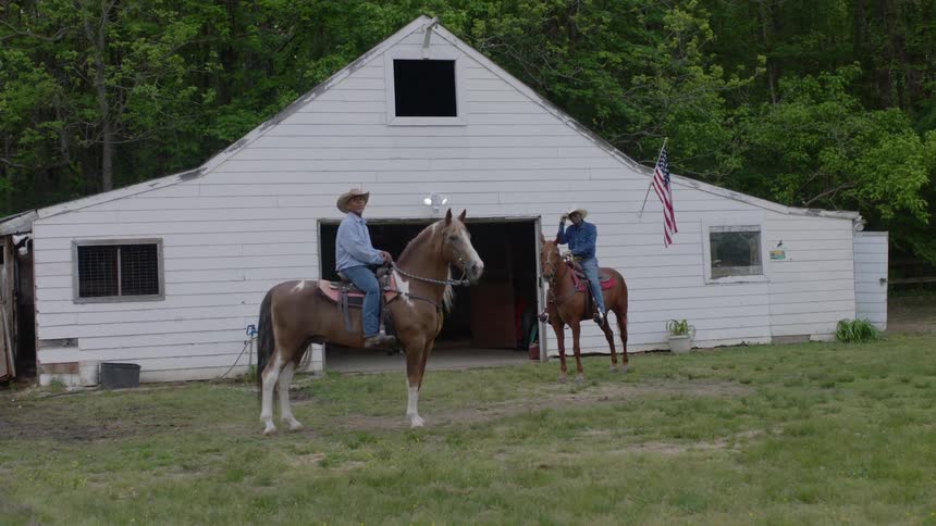 Two men wearing cowboy hats are sitting on horses outside of a barn flying an american flag