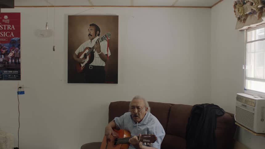 A man is inside a room sitting on a couch playing an acoustic guitar