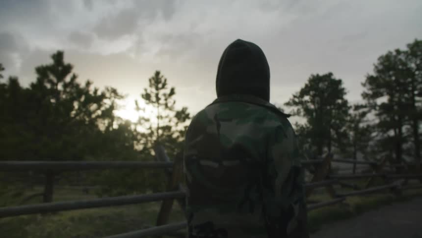 A person wearing a hoodie and camo jacket pauses to look at the sun low in the sky