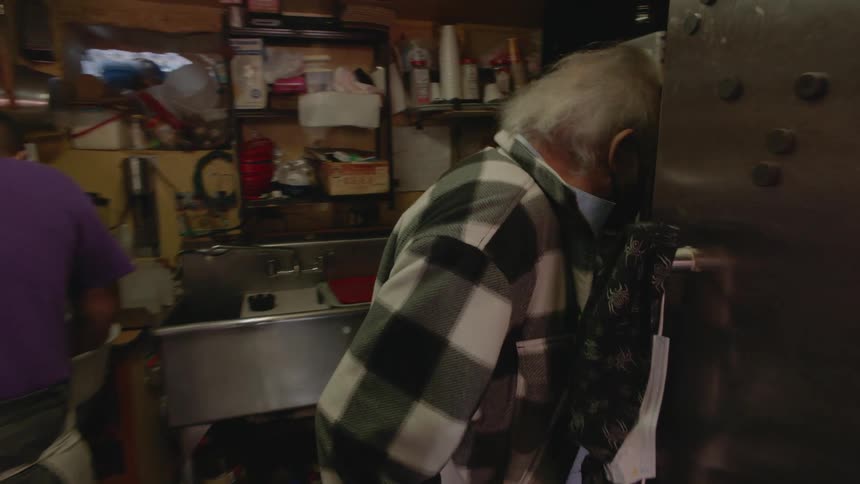 Older man wearing face mask walks through a very cramped kitchen where another man stands working