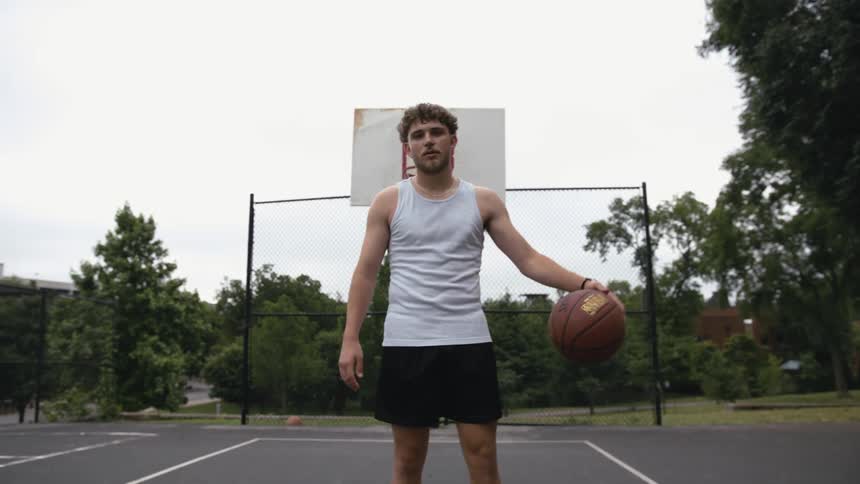 A man looks into the camera while dribbling a basketball on an outdoor basketball court on a cloudy day