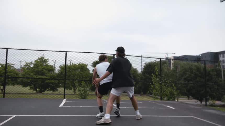 Two friends playing on a basketball court in a park one defending while the other takes a jump shot