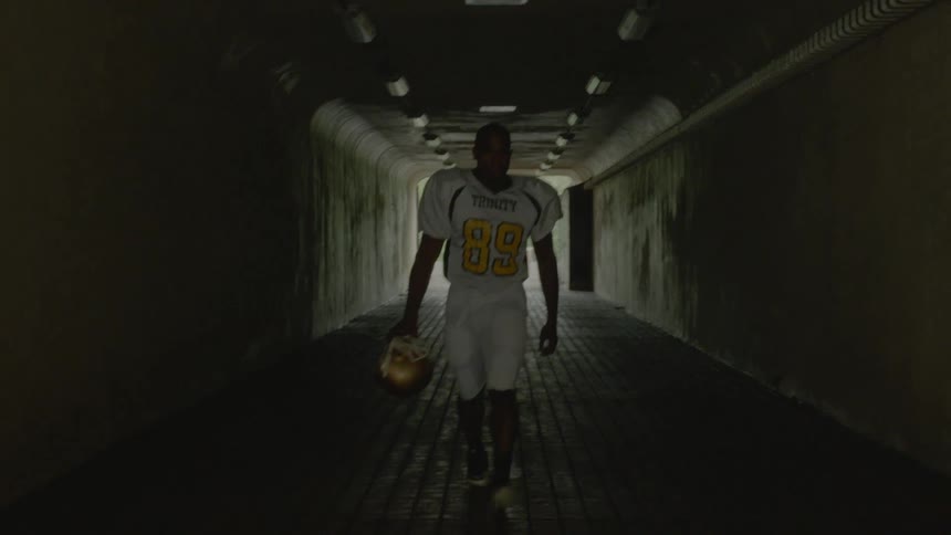 A young black man in a football uniform carrying a helmet walks through a tunnel