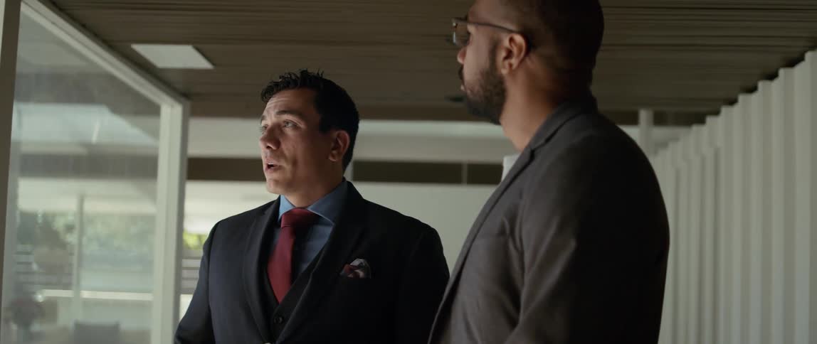 Two men are standing near a window and one man is looking around as the other man speaks to him