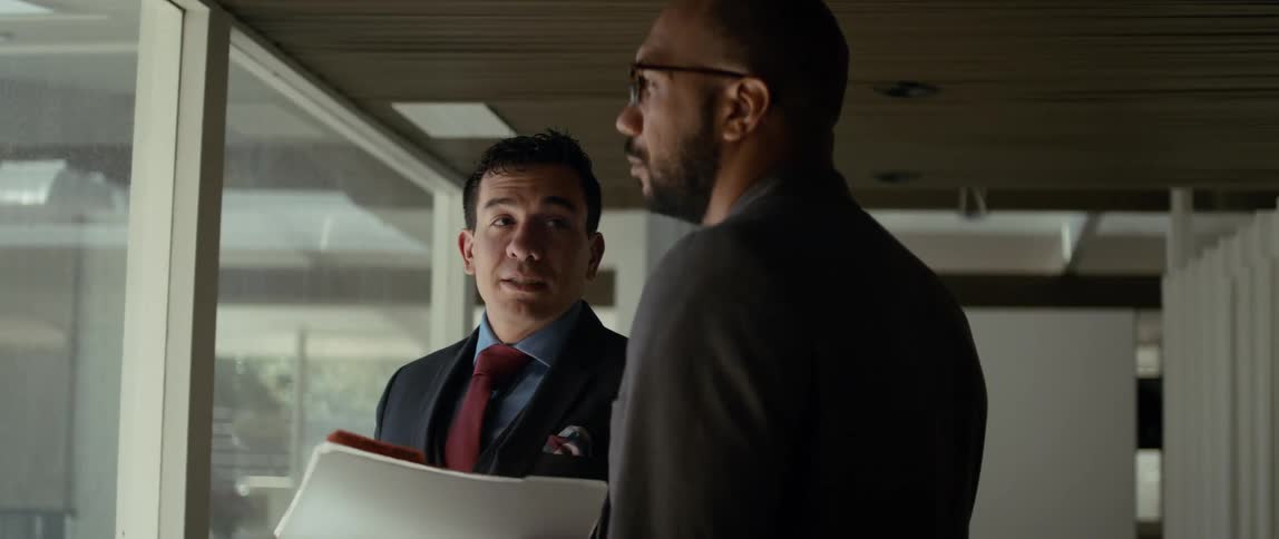 Two men are standing near a window and both move their hands as they look out the window and speak