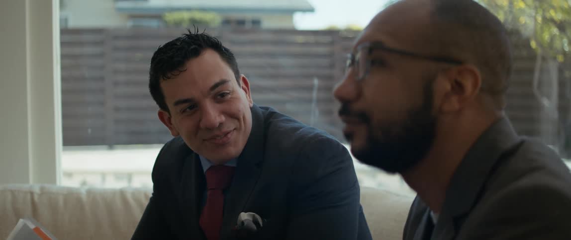A man in a business suit smiles at another sitting beside him