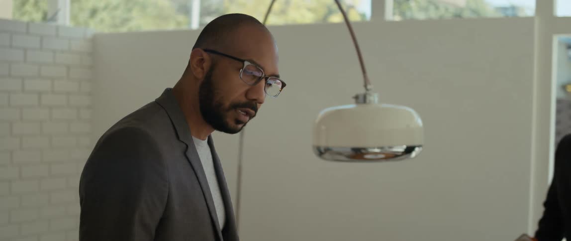 A man is walking in a room and looking around as another man sits down