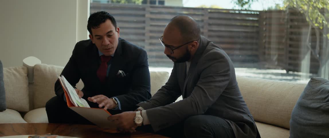 Two men are sitting on a couch looking at a notebook that one man is holding as he is speaking