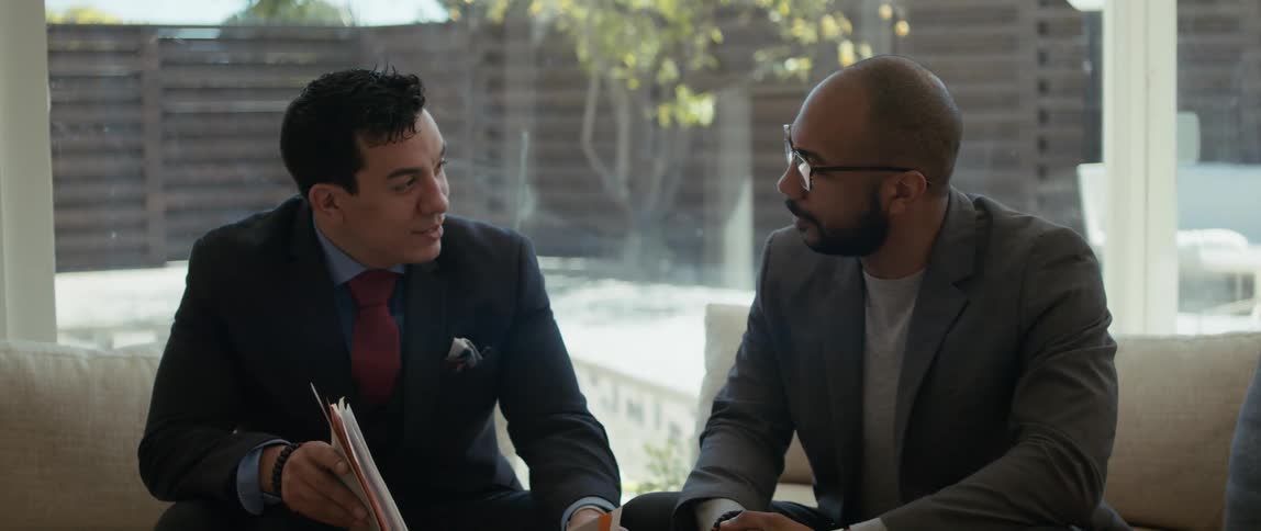 Two men sit in an office and talk as one of them shows the other some papers