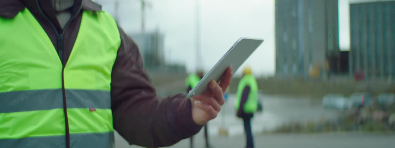 One person wearing a green vest holds a tablet while two people wearing vests stand behind him