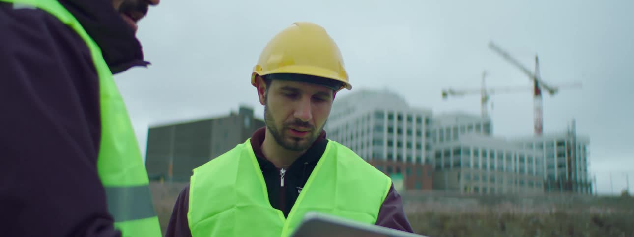 A construction worker nods while having a discussion with buildings and cranes behind him