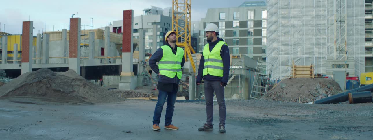 Two construction workers on a site bump fists and then talk with one another as one points in to the distance