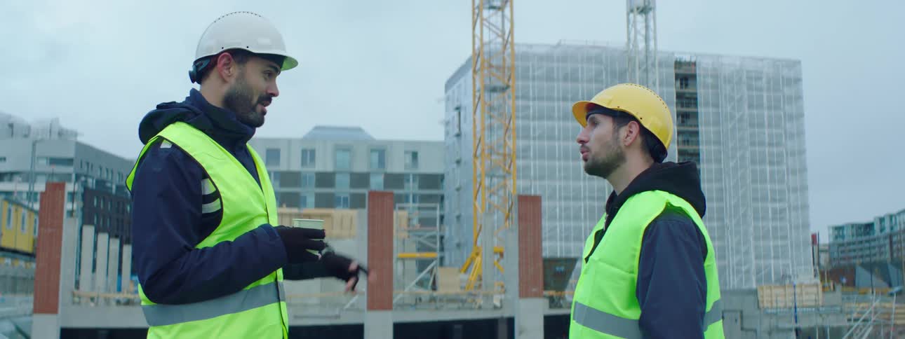 Two male construction workers stand and talk at a construction site in the city