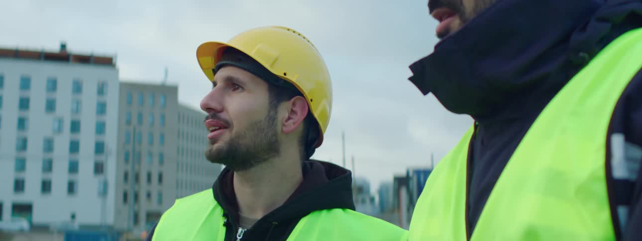 Two construction workers wearing safety vests talk as they stand outdoors