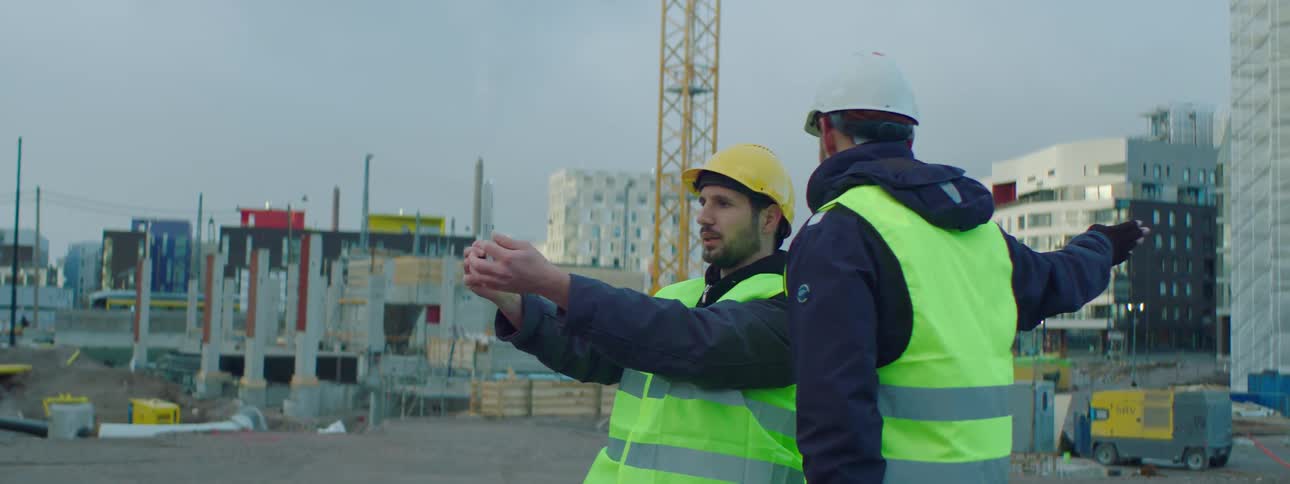 Two men outdoors on a construction site are using a tablet while discussing work on an cloudy day