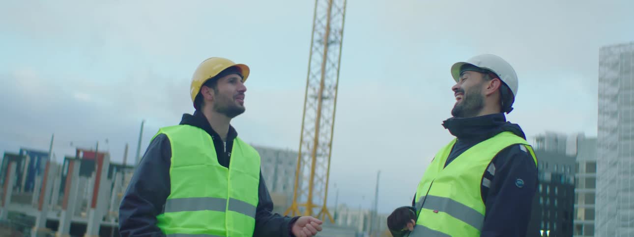 Two male construction workers wearing safety helmets and vests standing at a construction site talking and laughing together