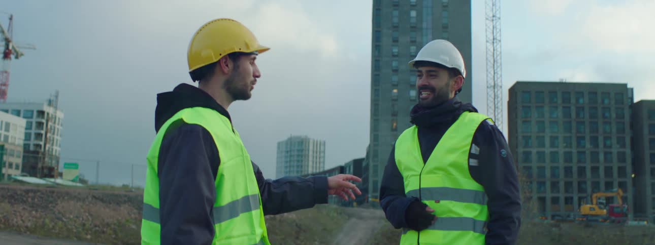 Two construction workers stand talking and laughing with cranes buildings and equipment behind them
