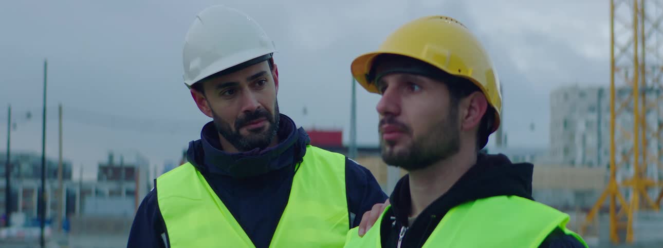 Male construction worker wearing a safety helmet and green vest standing at the construction site with a sad face while his friend approaches and touches his shoulder in a helpful and friendly way