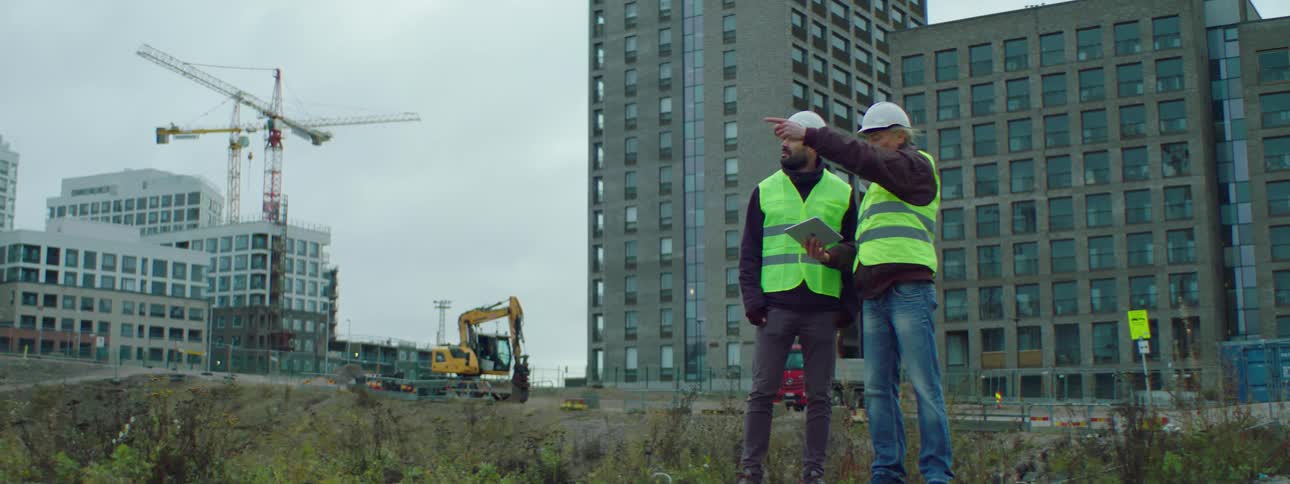 A man holding a tablet talks to a construction worker as he points off in the distance