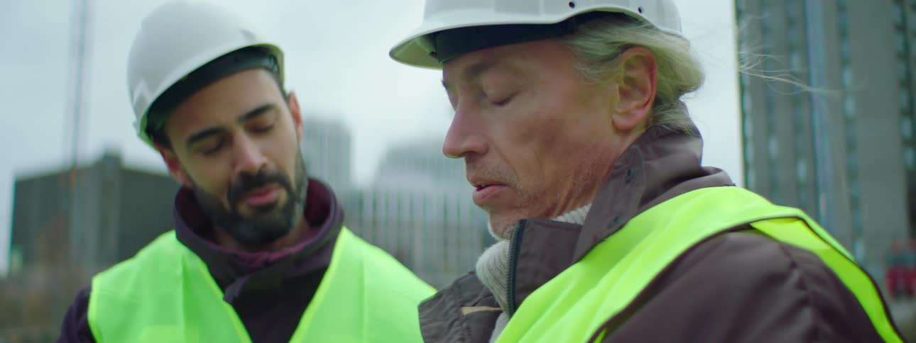 Two construction workers are standing outside looking at something and talking to each other